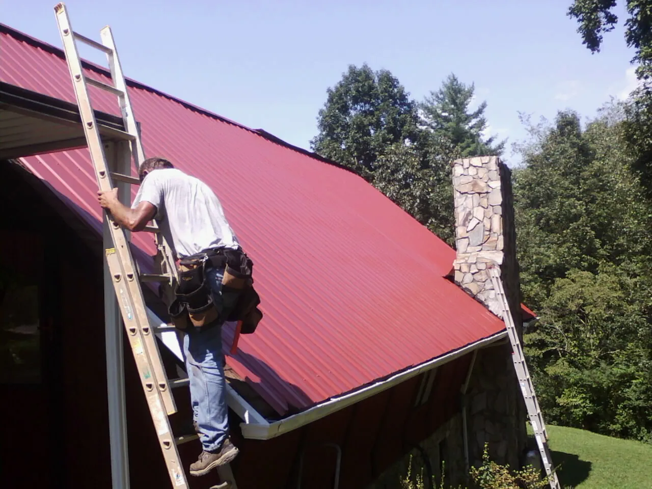 Roofer on a ladder beside a red metal roof and stone chimney — Soffit & Fascia Repair by Roofing Brothers in Lyons
